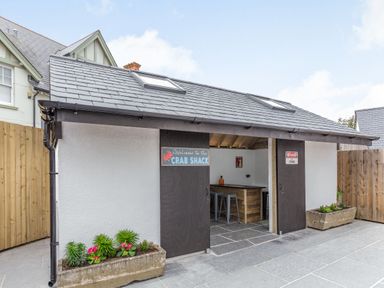 A small outdoor building with two doors and a sign reading Welcome to the Crab Shack with stools and a bar inside at The Old Coach House in Braunton