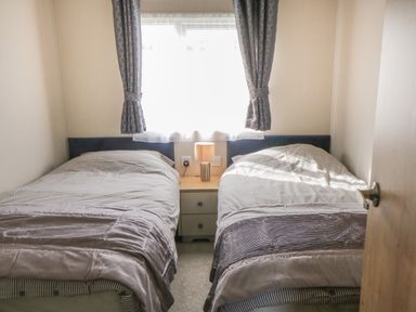 A bedroom with two single beds separated by a nightstand with a lamp and a window with curtains at Daisy Lodge in Cayton Bay near Cayton
