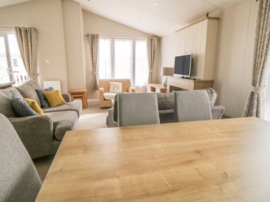 A living room with a wooden table gray chairs a gray sofa an armchair a television and curtains at Daisy Lodge in Cayton Bay near Cayton