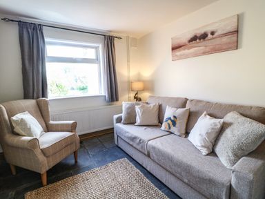 A living room with a grey sofa with pillows an armchair near a window and a lamp at Hops Cottage in Doveridge