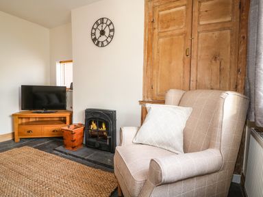 A living room with an armchair and a striped pillow next to a fireplace and a wooden cabinet at Hops Cottage in Doveridge