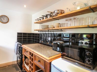 A kitchen corner with black tiled backsplash wooden shelves with pots pans plates glasses a microwave and toaster on counters at Hops Cottage in Doveridge