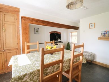 A dining area with a table covered by a green patterned tablecloth and wooden chairs near a wooden built-in cupboard and a kitchen with a stove at Hops Cottage in Doveridge