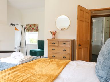 A bedroom with a wooden chest of drawers a round mirror a green chair and an open door leading to a bathroom at Hops Cottage in Doveridge