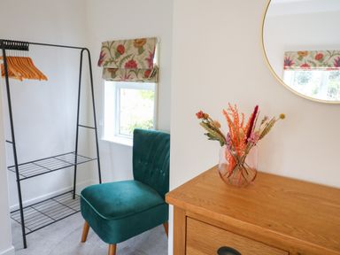 A corner of a room with a black metal clothing rack with wooden hangers a green upholstered chair a wooden dresser with a vase of dried flowers and a round mirror on the wall at Hops Cottage in Doveridge
