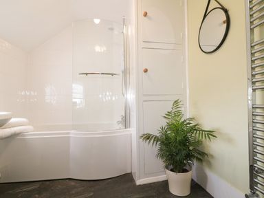 A bathroom with a curved bathtub and shower screen a round mirror a potted plant and built in storage cabinets at Hops Cottage in Doveridge