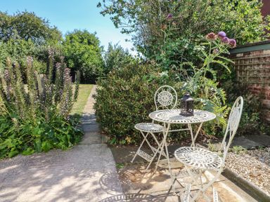 A garden with metal table and chairs on a paved area surrounded by plants and a lawn at Hops Cottage in Doveridge