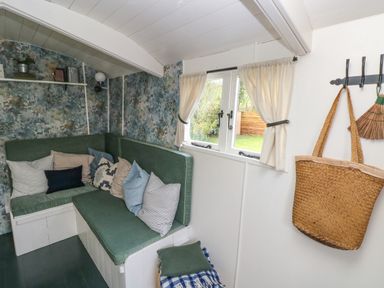 A seating area with green cushions and pillows next to a window with white curtains and a wall hook holding a woven bag and a broom at Hazel Nook in Narberth