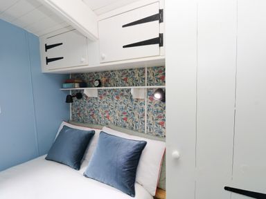 A bedroom with a bed with blue pillows under white cabinets and floral wallpaper at Hazel Nook in Narberth
