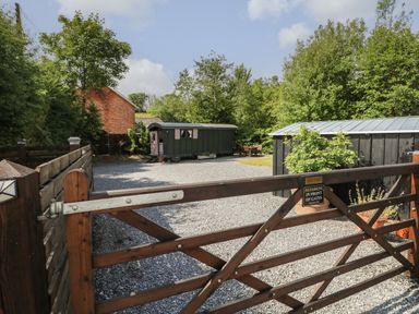 A gravel driveway with a wooden gate leading to a green shed and a black garage surrounded by trees and a brick building in the background at Hazel Nook in Narberth