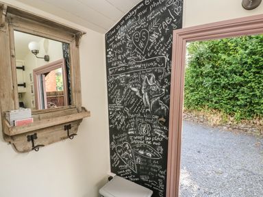An interior corner with a large wall mirror with a shelf and hooks a chalkboard wall with drawings and writings and an open door showing a gravel path and greenery outside at Hazel Nook in Narberth