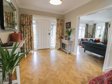 An entrance hall with wooden parquet flooring two tables with plants and decorative items a white front door and a view into a living room with sofas and windows at Pengwern in Llanrwst