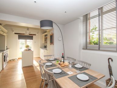 A dining area with a wooden table set with plates glasses and wine next to a kitchen with white cabinets and a window with blinds at St Mary's Chapel in West Lulworth