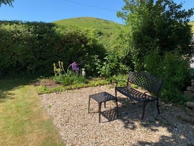 A garden area with a metal bench and table on a stone patio surrounded by grass and bushes at St Marys Chapel in West Lulworth