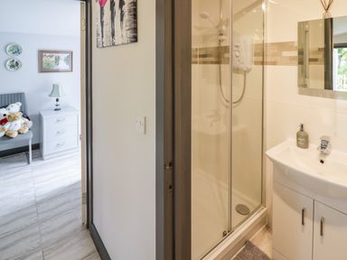 A shower with glass doors and a sink with a soap dispenser next to a room with a chair and teddy bears at The Garden House in Upper Quinton near Lower Quinton