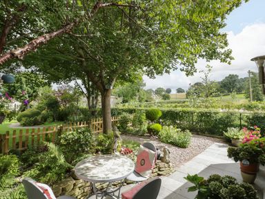 A garden patio with a table and chairs under a tree overlooking plants and a wooden fence at The Garden House in Upper Quinton near Lower Quinton
