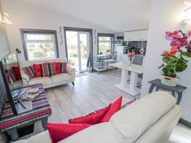 A living room with two white sofas with red and striped cushions a TV on a striped bench a dining table with flowers and a kitchen area at The Garden House in Upper Quinton near Lower Quinton