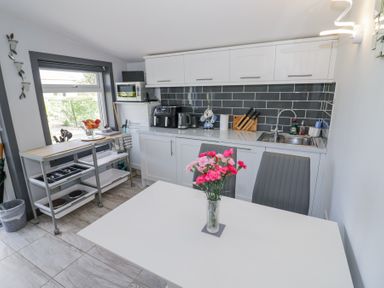 A kitchen with white cabinets a black tiled backsplash a sink a table with a vase of pink flowers and two gray chairs at The Garden House in Upper Quinton near Lower Quinton
