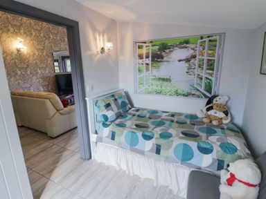 A small bedroom with a single bed covered with a geometric patterned blanket and three teddy bears on and near the bed a wall decoration resembling an open window with a garden view and a doorway showing a living room with a beige sofa and a television at The Garden House in Upper Quinton near Lower Quinton