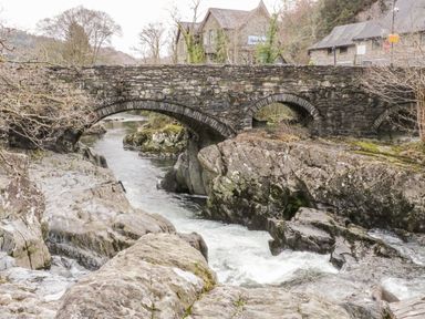 A stone arch bridge over a flowing river with rocky banks and some buildings in the background at Afallon in Penmachno