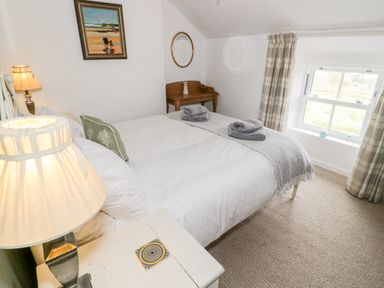 A bedroom with a bed with white linens and folded towels a bedside table with a lamp a small wooden desk with a mirror and a window with plaid curtains at Afallon in Penmachno