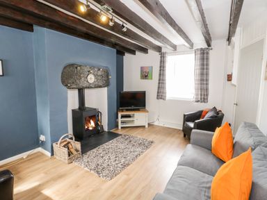 A living room with a grey sofa with orange cushions a black armchair a wood burning stove a television on a stand and exposed ceiling beams at Afallon in Penmachno