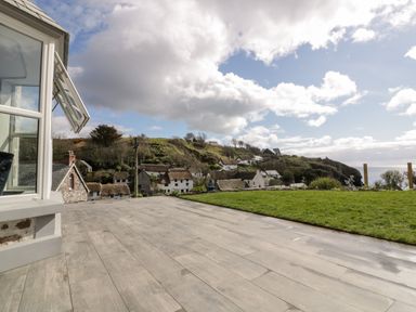 A patio with wooden flooring and a grassy area overlooking houses on a hillside by the sea at Louvain in Cadgwith near Ruan Minor