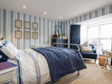 A bedroom with a bed covered with blue and white bedding a blue armchair a chest of drawers and striped wallpaper at Louvain in Cadgwith near Ruan Minor
