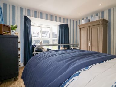 A bedroom with a bed covered in a blue duvet a wardrobe a chest of drawers and a window with blue curtains at Louvain in Cadgwith near Ruan Minor