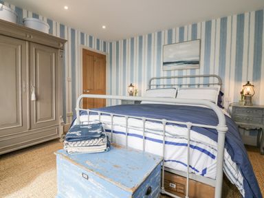 A bedroom with a metal bed frame bedding a distressed blue chest folded blankets a wooden wardrobe and striped wallpaper at Louvain in Cadgwith near Ruan Minor