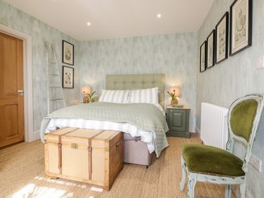A bedroom with a bed, two bedside tables with lamps, a wooden chest at the foot of the bed, framed pictures on the walls, a green chair, and a wooden door at Louvain in Cadgwith near Ruan Minor