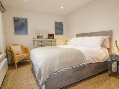 A bedroom with a bed, wicker chair, dresser with mirror, two framed pictures, and flowers at Louvain in Cadgwith near Ruan Minor