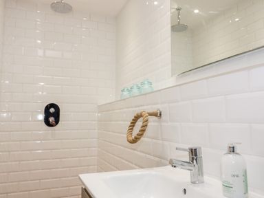 A bathroom with white tiled walls a sink a faucet a mirror a soap dispenser glass jars and a showerhead at Louvain in Cadgwith near Ruan Minor