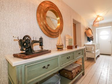 A hallway with a green sideboard holding an old sewing machine model boat and decorative boxes with a round mirror above and a bench near the door at Louvain Cadgwith near Ruan Minor