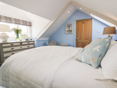 A bedroom with a white bed and a blue pillow with birds a dresser with a lamp flowers and a record player on top and a wooden door at Louvain in Cadgwith near Ruan Minor