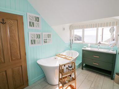 A bathroom with a white freestanding bathtub and wooden storage rack next to a green vanity under windows at Louvain in Cadgwith near Ruan Minor