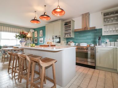 A kitchen with a central island and wooden stools a dining table and teal tiled backsplash at Louvain in Cadgwith near Ruan Minor