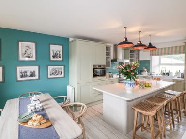 A kitchen with an island and wooden stools next to a dining table with plates and cups on a teal wall at Louvain Cadgwith near Ruan Minor