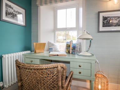 A desk with a lamp an open book and magazines by a window with a wicker chair in a room with framed photos on the walls at Louvain Cadgwith near Ruan Minor