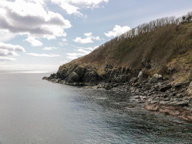 A rocky coastline with a hill covered in sparse trees and shrubs overlooking the sea at Louvain in Cadgwith near Ruan Minor