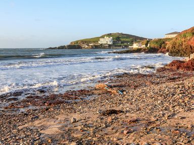 A rocky beach with waves and buildings on a hill in the background at 2 The Bay in Bigbury