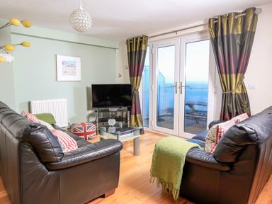 A living room with two black leather sofas a glass coffee table a television and double glass doors with striped curtains at 2 The Bay in Bigbury