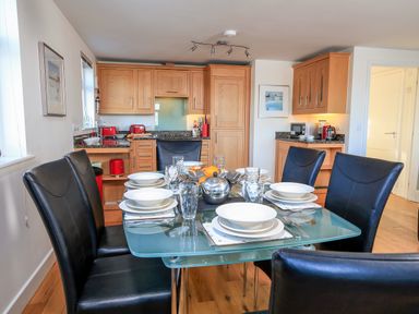 A dining area with a glass table set for six and black chairs in a kitchen with wooden cabinets at 2 The Bay in Bigbury