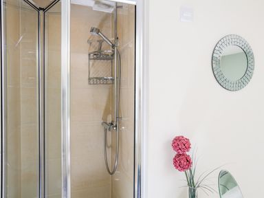 A bathroom with a glass-enclosed shower, a round decorative mirror on the wall, and a vase with pink flowers at 2 The Bay in Bigbury
