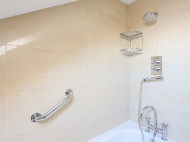 A bathroom shower area with beige wall tiles a chrome grab bar a showerhead a two tier metal corner shelf and a bathtub faucet at 2 The Bay in Bigbury
