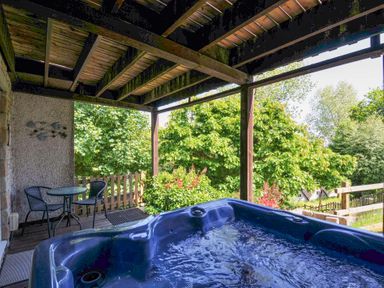 A covered outdoor deck with a hot tub and a small table with two chairs surrounded by trees at 59 Valley Lodge in Callington near Gunnislake