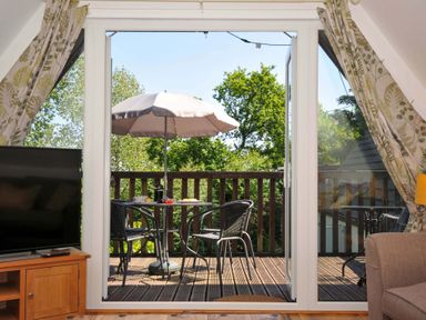 A balcony with a table umbrella and two chairs viewed from a room with a television and curtains at 59 Valley Lodge Callington near Gunnislake