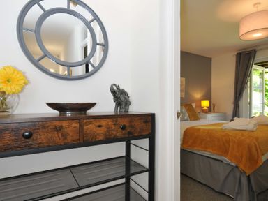 A hallway with a wooden console table and round mirror leading to a bedroom with a bed and a bedside lamp at 59 Valley Lodge in Callington near Gunnislake