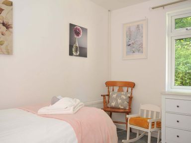 A bedroom corner with a bed covered in white and pink bedding two chairs a wooden chair with a cushion a white rocking chair with an orange blanket a white chest of drawers and three framed pictures on the walls at 59 Valley Lodge Callington near Gunnislake