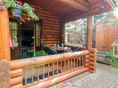 An outdoor wooden deck with a round glass table and four chairs next to a log cabin with an open door and a small wooden shed in the background at Jalan End in Felton
